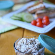 A small bowl filled with the meat salad. There is a plate with a sandwich with vegetables in the background