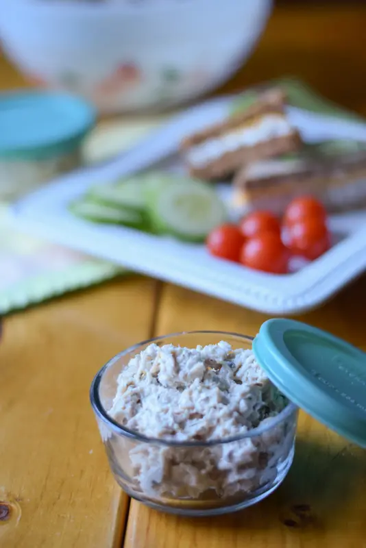 A small bowl filled with the meat salad. There is a plate with a sandwich with vegetables in the background