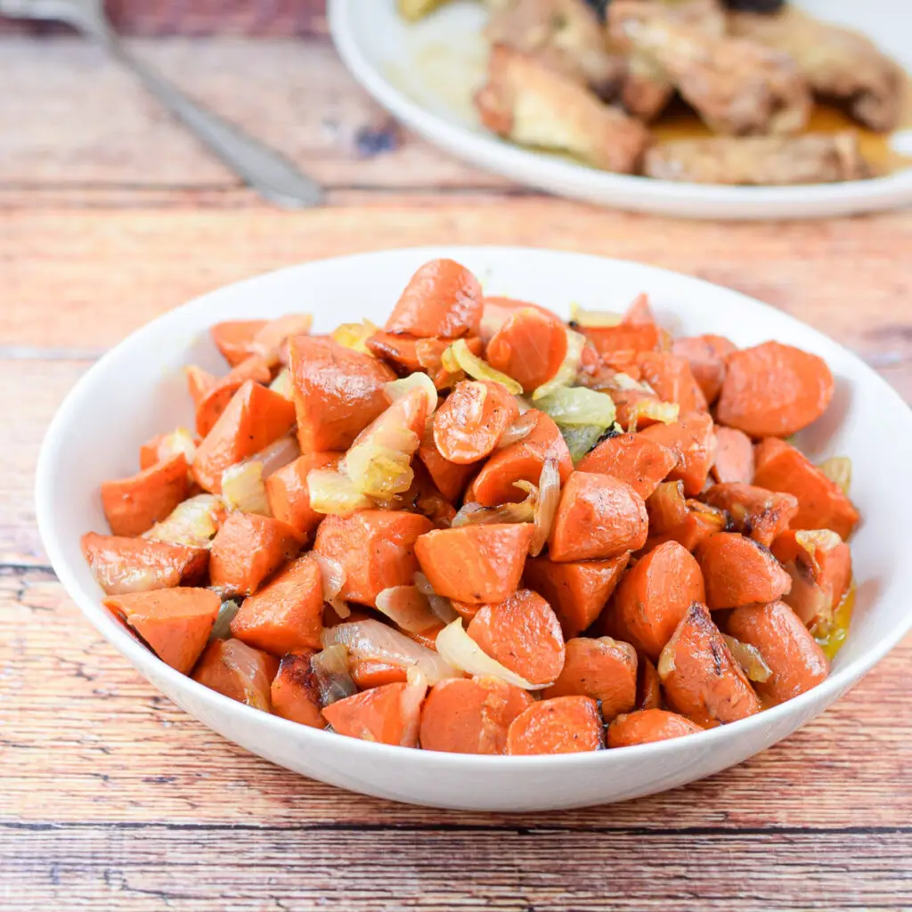A white bowl of carrots and onions in front of a plate with chicken wings, potatoes and a salad in a bowl - square