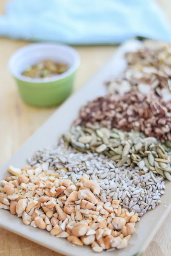 A bunch of nuts and seeds on a rectangle platter and a ramken in the background