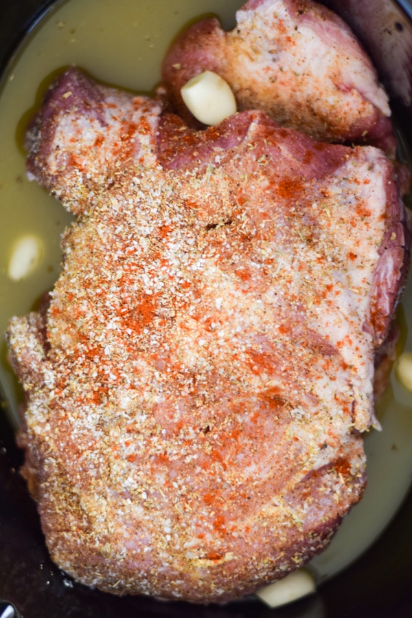 Pork shoulder in the insert for the crockpot with the rub on it, garlic cloves and broth