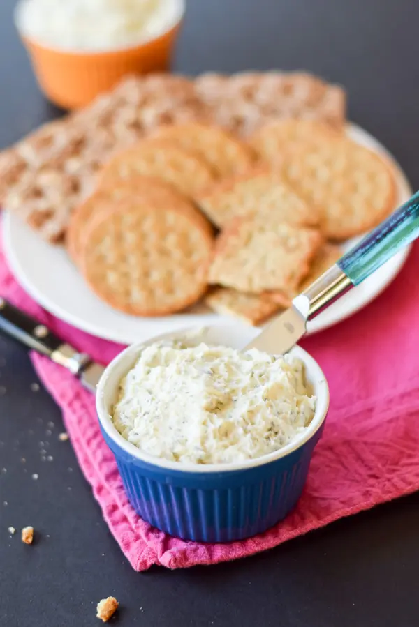A blue bowl filled with the buttery spread with crackers in the background on a plate