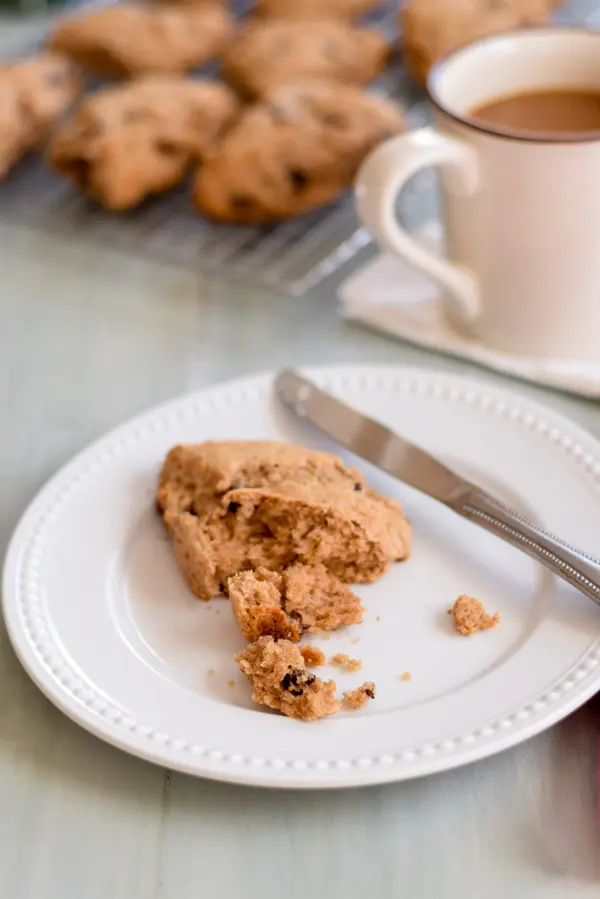 A scone on a white beaded plate with coffee and more scones in the background