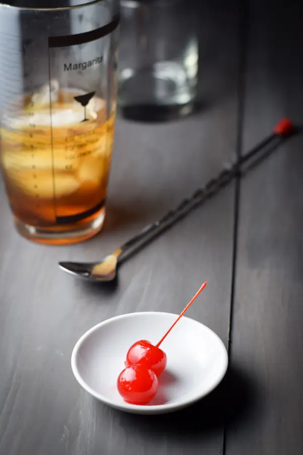 Two cherries on a small white dish with the spoon, shaker and glass in the background