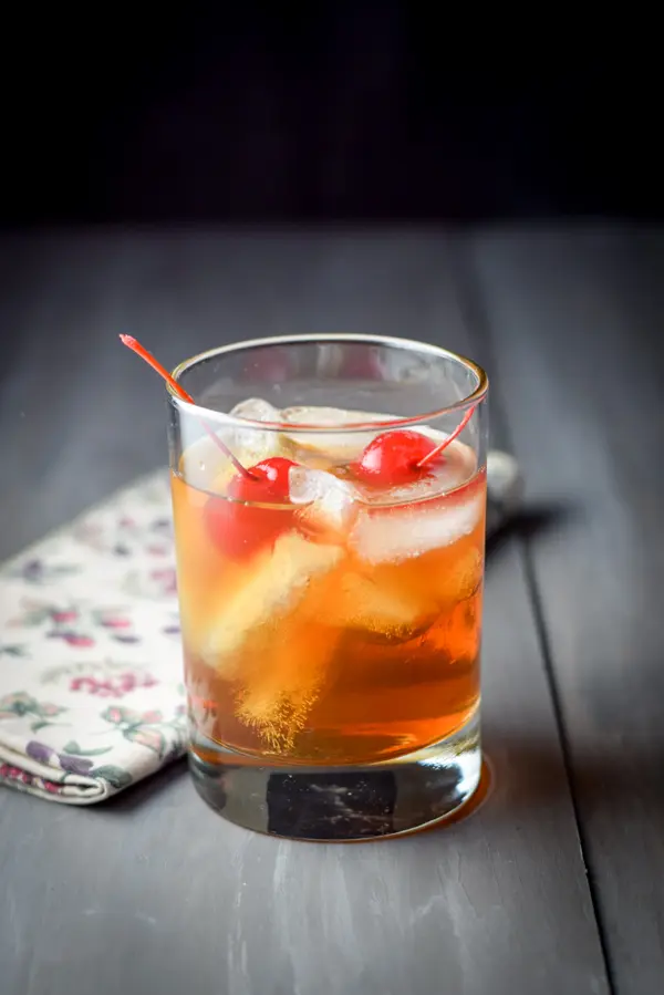 A double old fashioned glass with the whiskey cocktail with two cherries in it