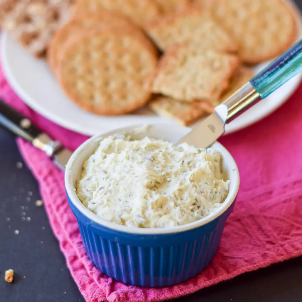 A blue bowl filled with the buttery spread with crackers in the background on a plate - square