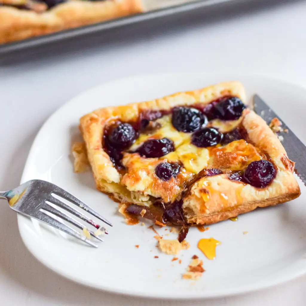 A white plate with flakey puff pastry covered with cherries, onion and cheese in front of the pan of tarts - square