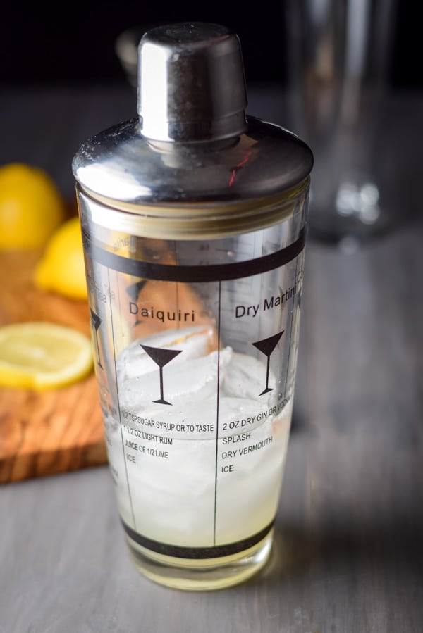 Shaker is capped and ready for it to be shaken. There is a board with lemon slices and a glass in the background