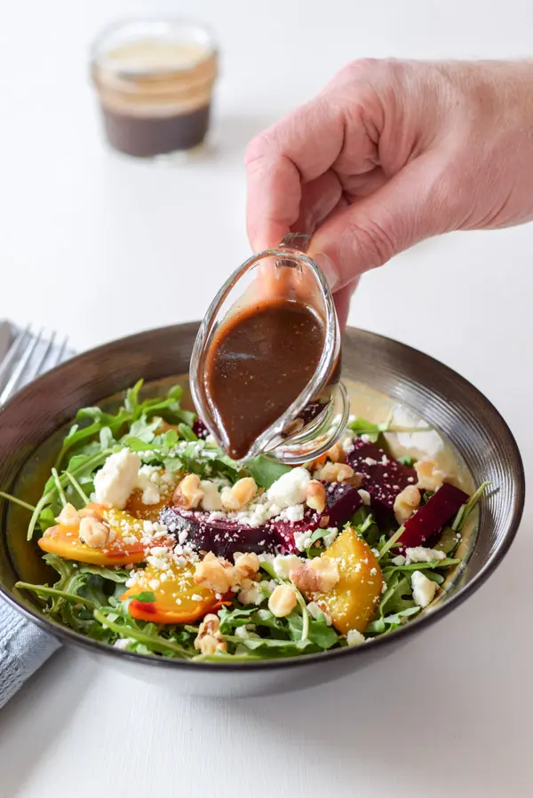Hand holding small pitcher of balsamic vinaigrette dressing over the salad, ready to pour.