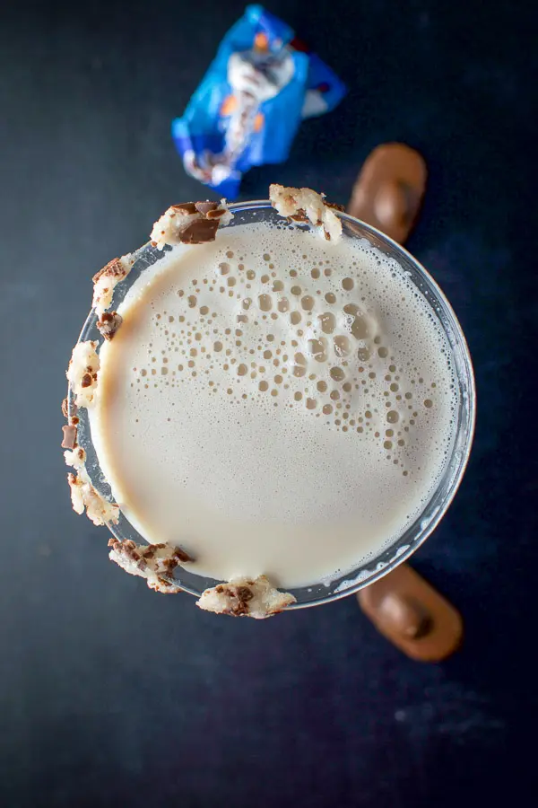 Overhead view of the the milky cocktail with almond joy bars smooshed on the side of the glass. There are candy on the board and a wrapper