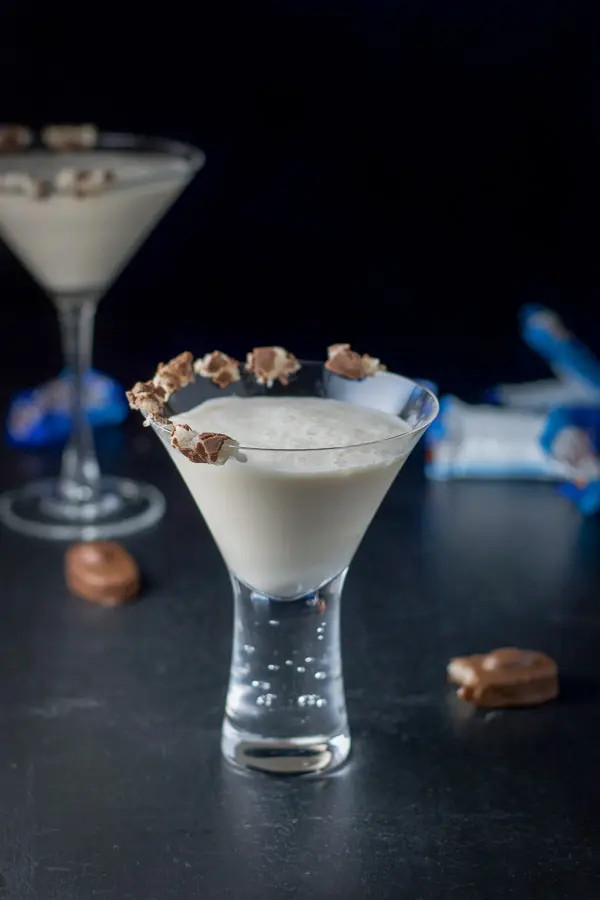 A milk cocktail in a bubble martini glass with the almond joy candy on the rim of the glass. There are candies on the table and wrappers in the background