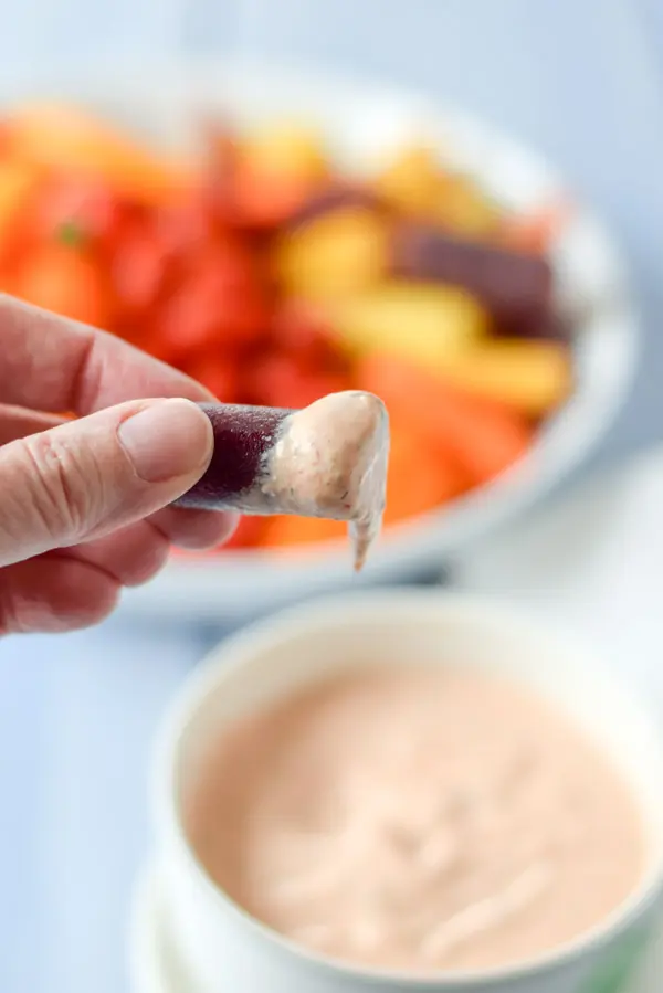 A hand holding a carrot dipped into the dip held over the bowl of dip. You can see the vegetables in the background