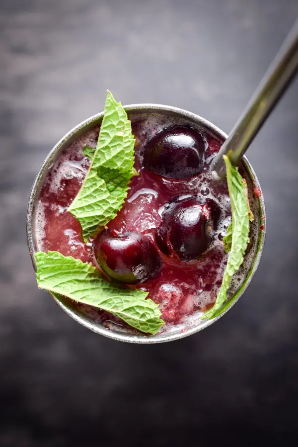 Overhead view of a metal glass with a straw. In the glass is liquid, ice, three cherries and mint