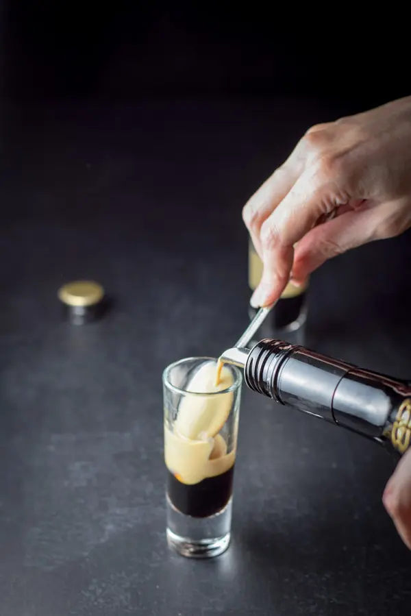 A female hand pouring Bailys Irish Cream over the back of a spoon into a shot glass that has kahlua in it already
