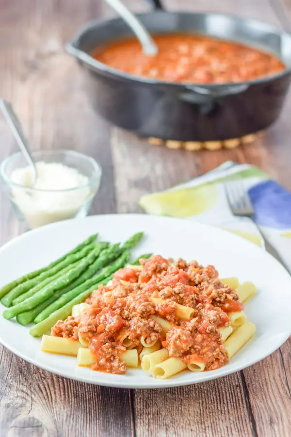 A white plate with a meat red sauce on pasta with asparagus and a pan of the gravy in the background