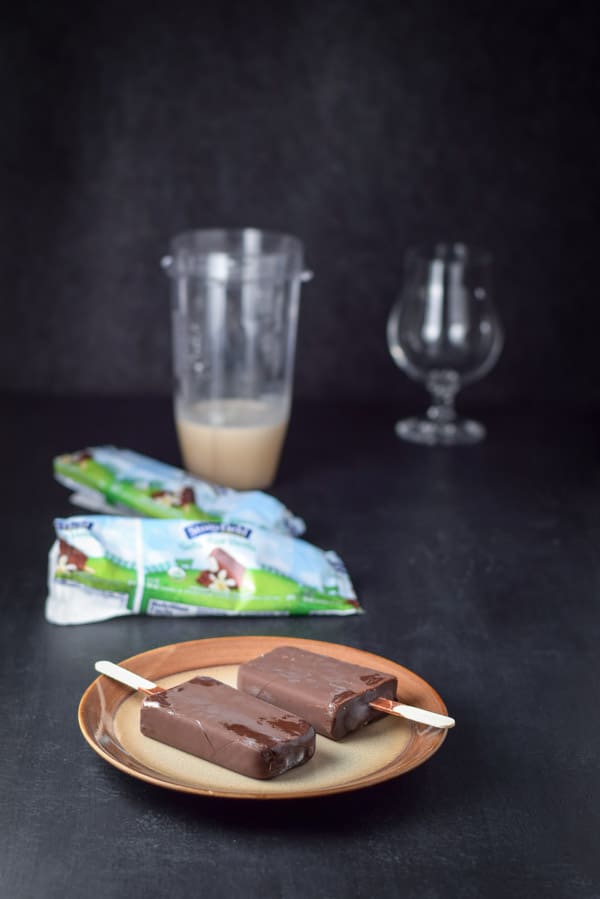 Two chocolate covered yogurt bars on a plate with the wrappers, blender container and glassware in the background