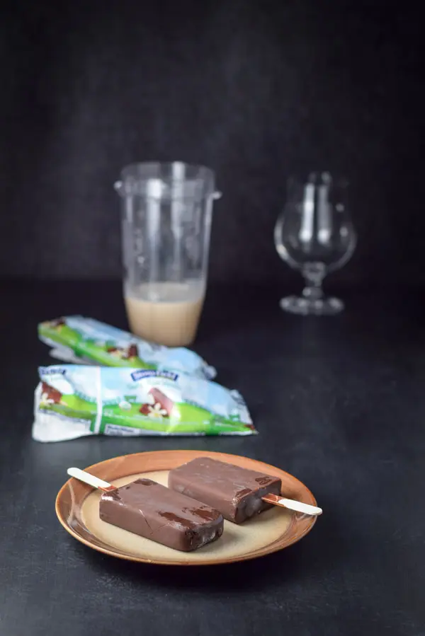 Two chocolate covered yogurt bars on a plate with the wrappers, blender container and glassware in the background