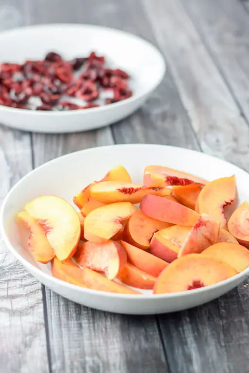 Two white deep bowls with sliced peaches in one and cherries in the other