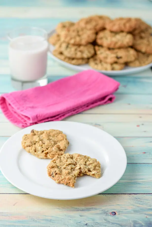 Two cookies on a white plate, one with a bite taken out of it with a big plate of piled up cookies and a glass of milk and napkin