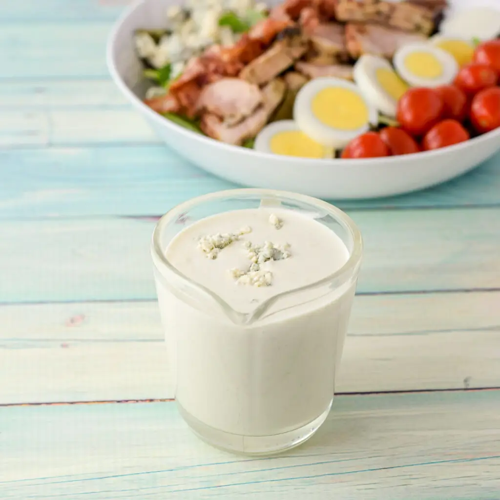 Dressing with blue cheese crumbled on top in a clear glass pitcher with two salads in the background - square