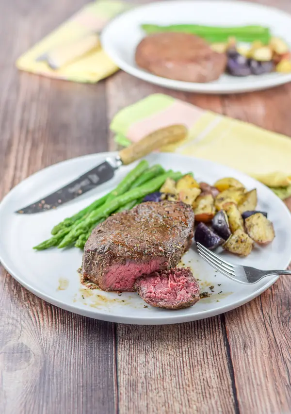 A cut piece of steak on the plate with potatoes, asparagus and knife and fork. there is another plate in the background