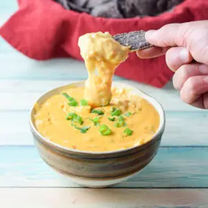 A hand holding a chip dripping cheesy dip into a bowl with chips in the background - square