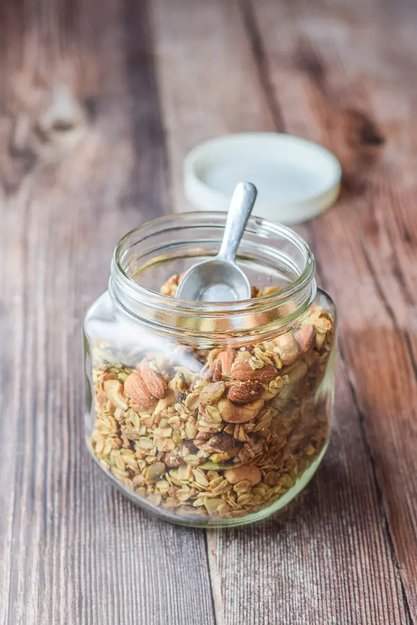 A large glass jar filled with nuts and granola with a small scoop in it