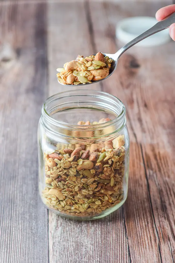 A hand holding a spoon of granola over the jar