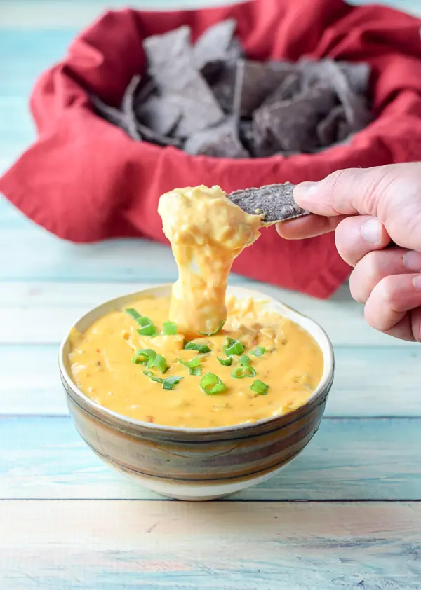 A hand holding a chip dripping cheesy dip into a bowl with chips in the background