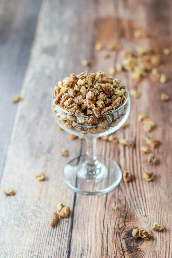 A coupe glass with the candied pecans in them and more pecans strewn across the wooden table