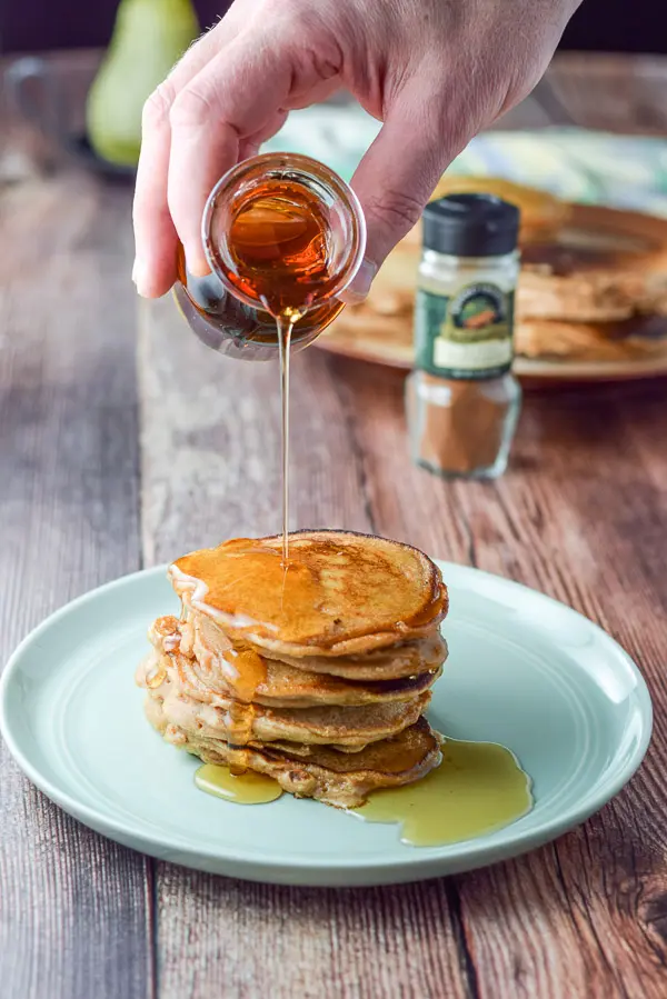 A hand holding a small glass bottle of maple syrup and being poured onto some pancakes. There is cinnamon and more pancakes in the background