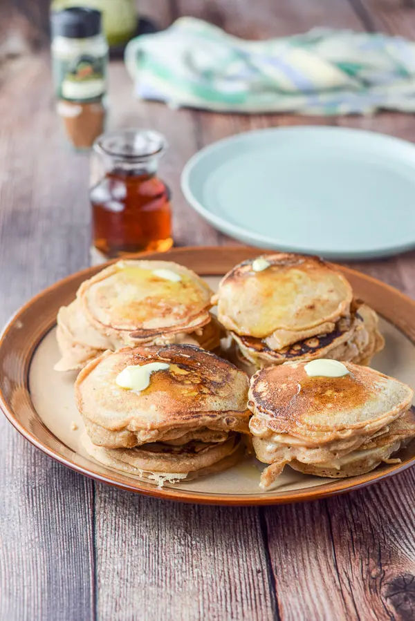 A brown plate with 4 stacks of pancakes and butter on them. There is a green plate, maple syrup and spices in the background