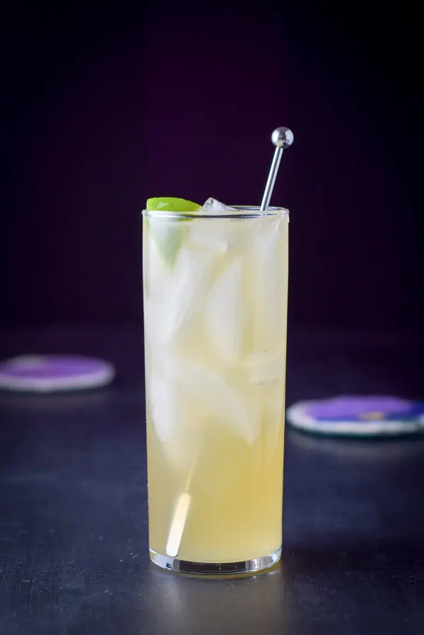 Vertical view of a collin glass with the yellow cocktail with a lime wedge and stirrer in it