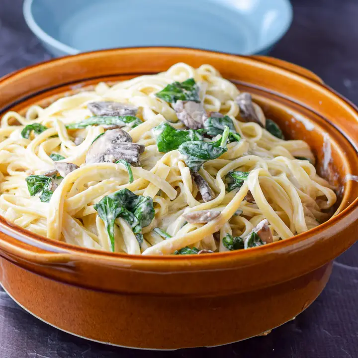 A big brown bowl of fettuccine with mushrooms and spinach with two bowls in the background - Square