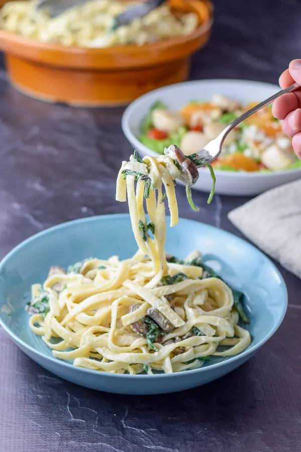 A forkful of mushroom and fettuccine held above the shallow bowl with a salad and the big bowl in the background