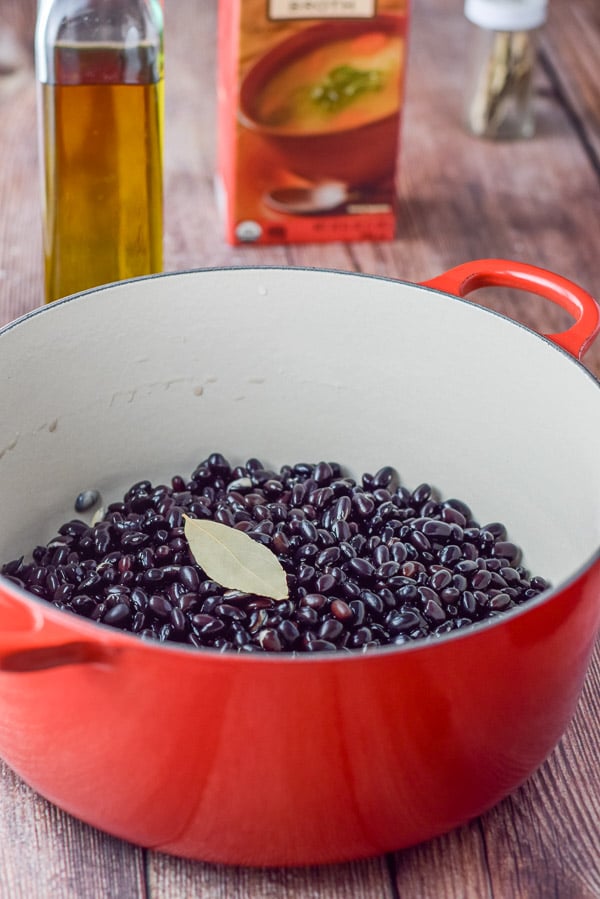 Black beans soaked over night and in a red pan. There is a bay leaf on them and broth and oil in the background