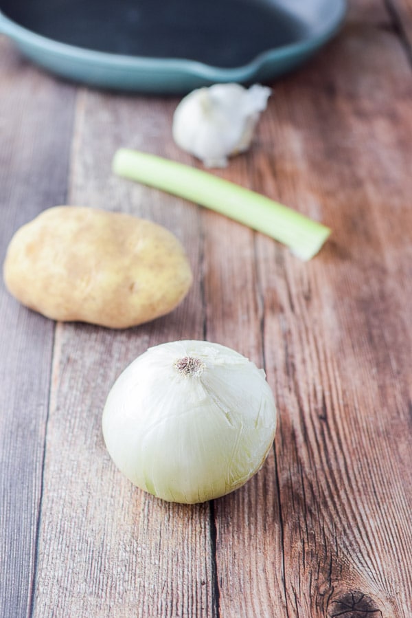 Onion, potato, celery and garlic on a table with a saut&egrave; pan in the background