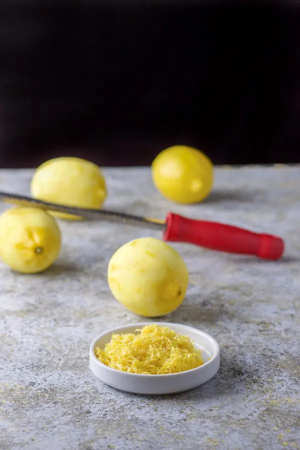 Lemon zest in a white dish in front of lemons and a microplane