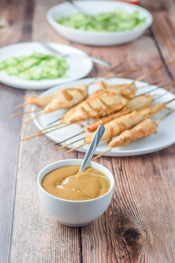 A white small bowl with sauce in it with a plate of chicken satay in the background