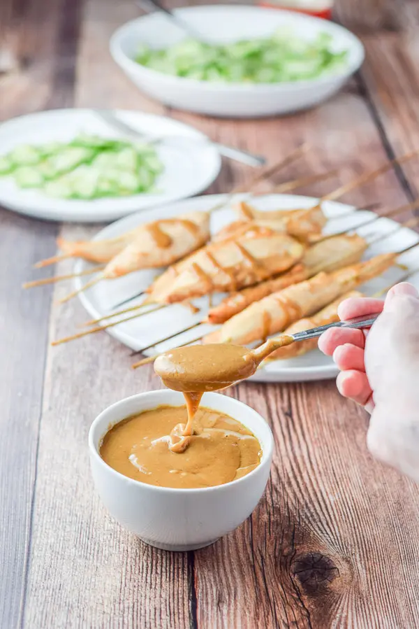 A hand holding a spoonful of sauce over the little bowl. There are chicken satay and a cucumber salad in the background