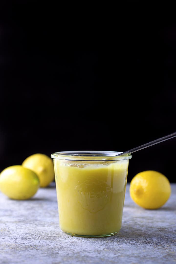 Vertical view of a jar of curd with a spoon in it and three lemons behind it