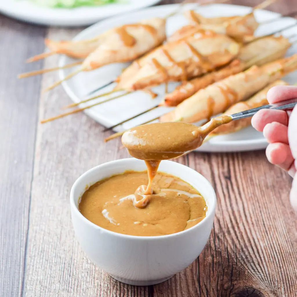 A hand holding a spoonful of sauce over the little bowl. There are chicken satay in the background - square