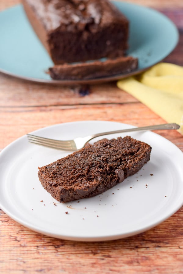 A slice of chocolate bread on a white plate with the loaf in the background