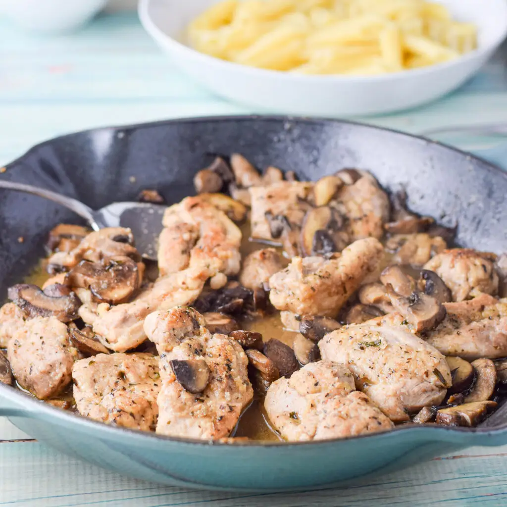 Chicken and mushrooms in the pan with a shallow bowl of pasta in the background - square