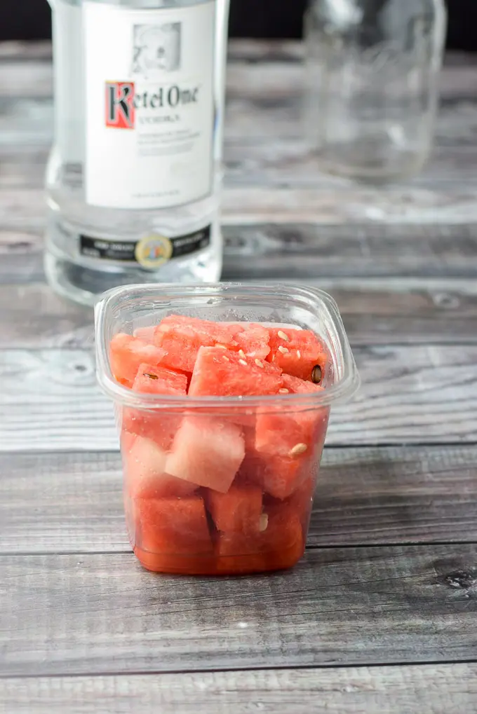 Container of watermelon, bottle of vodka and jar on a grey table