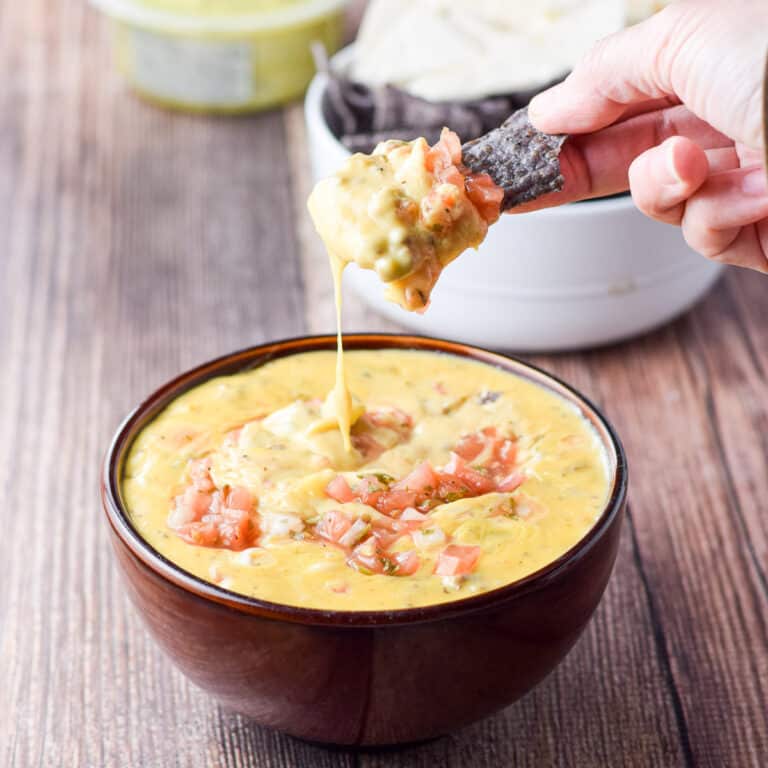 A hand holding a dripping chip of cheesy dip with guac, pita and chips in the background - square