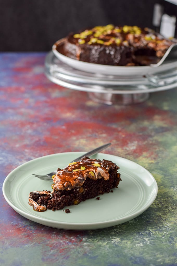 A piece of chocolate cake on a green plate with the cake on the pedestal in the background