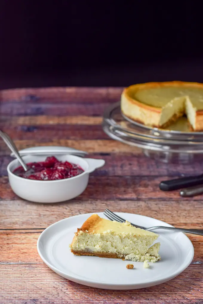 A piece of cheesecake on a plate with cherry sauce in a bowl and the cake on the pedestal