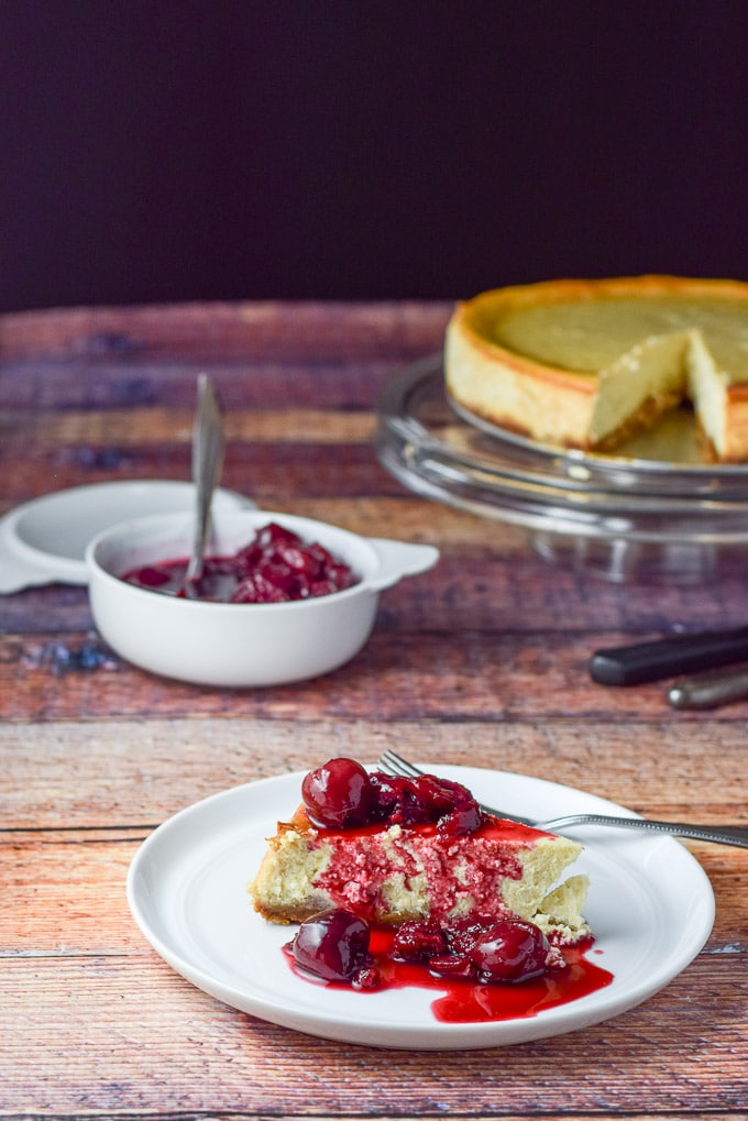 Cherry sauce poured on the cheesecake with the pedestal and bowl of sauce in the background