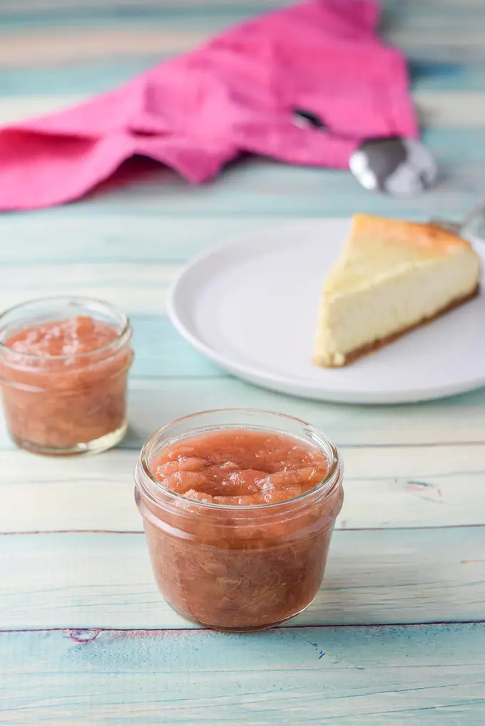 Two small Ball jars full of rhubarb spread in front of a plain cheesecake